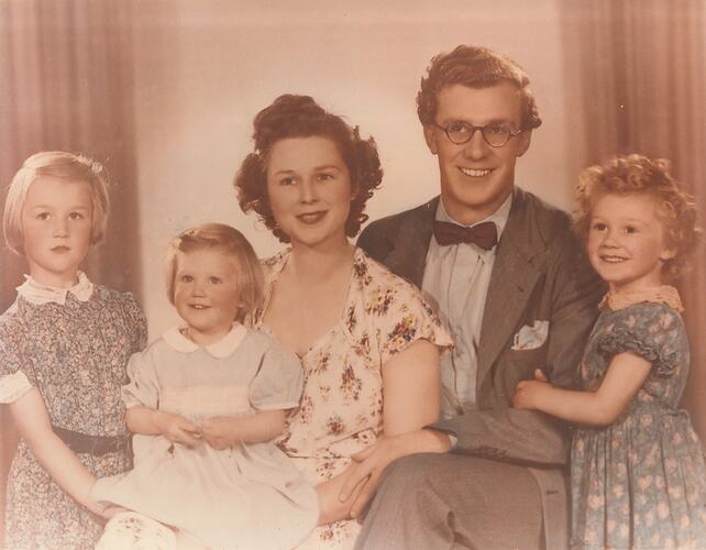 Man, woman with three young girls, indoors. He wears glasses and a bow tie, the woman and girls wear dresses.