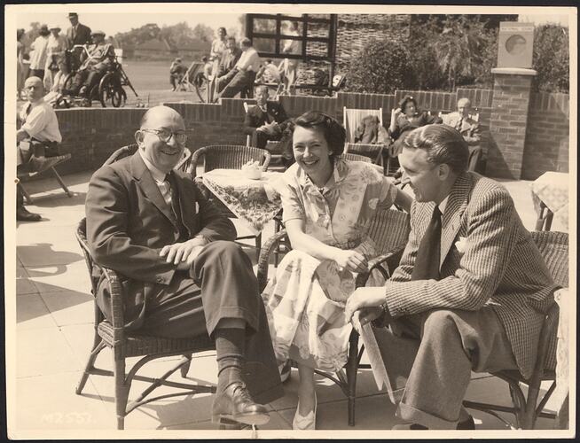 Smiling group seated in wicker chairs outdoors. Woman in between two men. More groups of people in background.