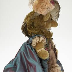 Profile of brown plush bear wearing lavish cream hat with feather and ornate dusty pink dress and blue jacket.