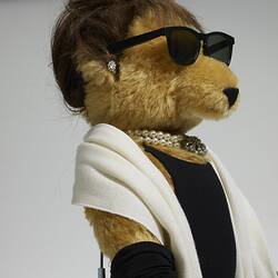 Detail of light brown bear. Wears black hat, sunglasses, dress, full-length gloves, white pearls and scarf.
