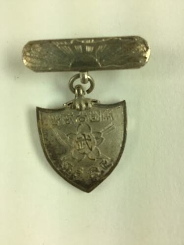 Decorative shield shaped medal suspended from bar pin.