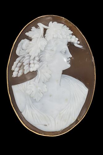 Oval brooch, white cameo of classical female facing right with a headpiece of grapes, leaves and feathers. Brown background set in polished gold border.