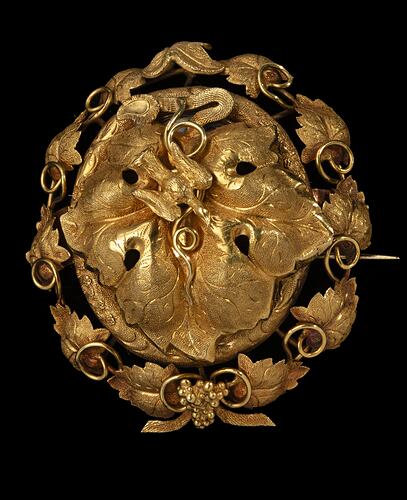 Gold oval locket brooch. Large central vine leaf with flying bird. Oval border surrounded by vine leaves and tendrils. Small bunch of grapes at base.