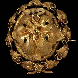 Gold oval locket brooch. Large central vine leaf with flying bird. Oval border surrounded by vine leaves and tendrils. Small bunch of grapes at base.