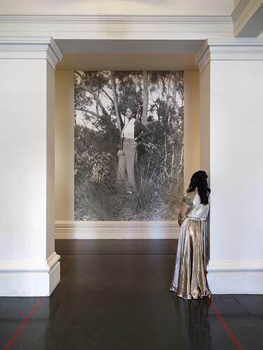View from behind of a woman in long dress leaning against an alcove wall viewing an historical photograph of a woman.