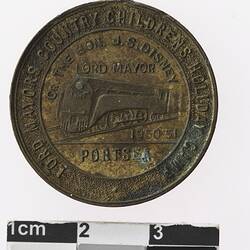 Round bronze coloured medal with train and text surrounding, rusted on surface.