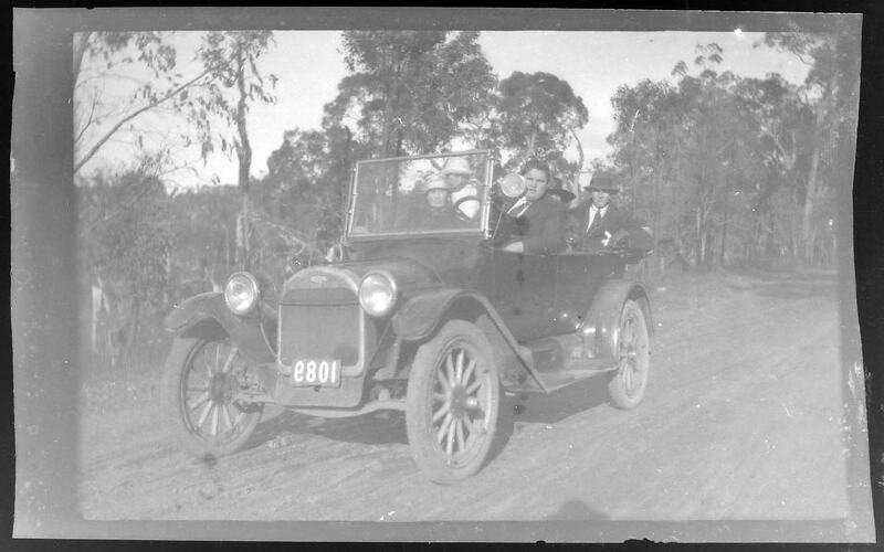 Men and women open-top motor car on tree-lined, dirt road.