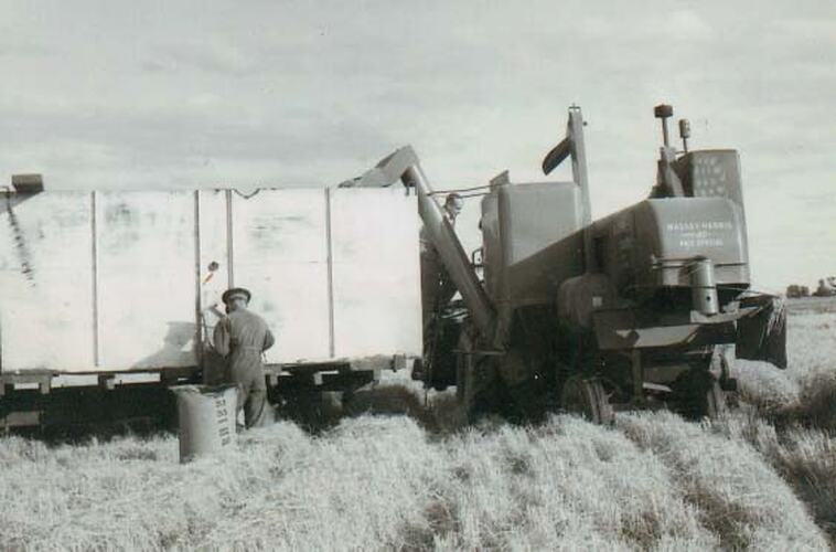 2 menoperating a harvester, showing grain bin and bag of rice in rice field.