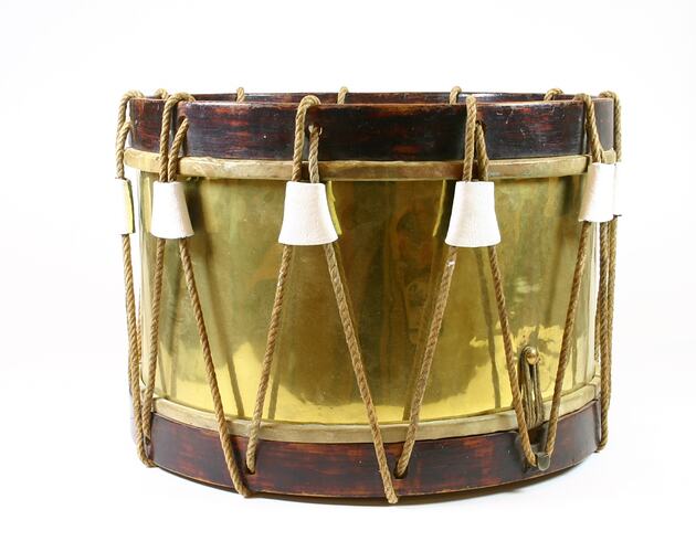 Cylindrical shiny brass drum, wooden edges. Rope threaded from top to lower edge.