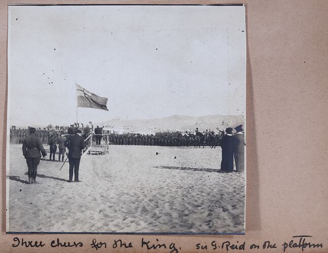 Soldiers standing in formation in front of platform with flag, onlookers watch on, handwritten text below.