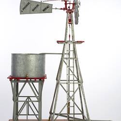 Model water pumping windmill next to water tank on a stand. Vanes side on facing right.