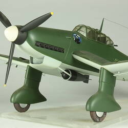 Detail of model aeroplane front and cockpit. Dark green, pale blue underside. Black propeller with white tip.