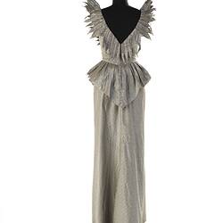 Evening dress, full length, silver-grey silk. No sleeves, winged shoulder peaks, ruffle at waist. Back view.