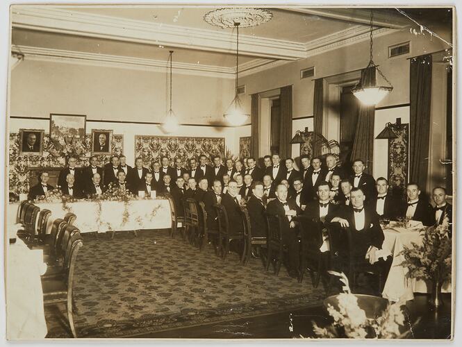 Large group of men, dressed in tuxedos, seated and standing around dining tables in horseshoe shape.