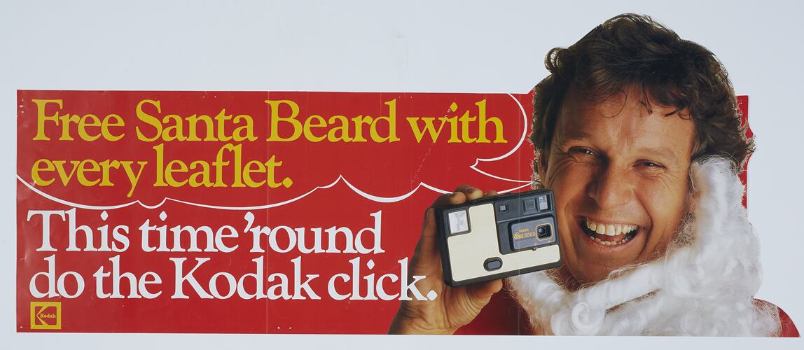 Man smiling and wearing a white Santa beard below his chin while holding up a camera to face level. Text at le