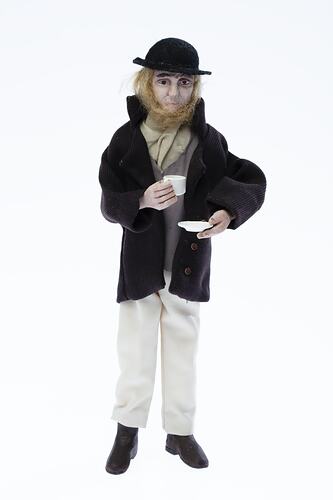Miniature man in dark coat, light pants. Holds a cup and saucer. Has a hat and fair hair.
