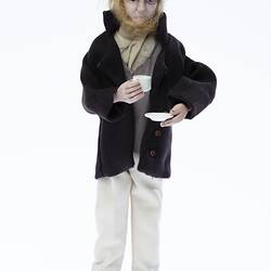 Miniature man in dark coat, light pants. Holds a cup and saucer. Has a hat and fair hair.