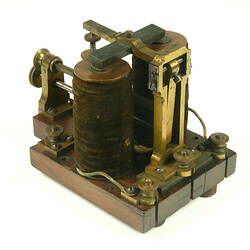 Brass apparatus with battery in centre on wooden base, three quarter view.