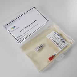 Labelled syringe and vial in lined plastic box on angle. Card in lid has printed text. White background.