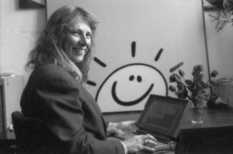 Black and white image of a woman sitting at a desk with a laptop on her knee.