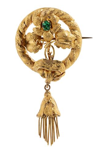 Round gold brooch. Top central flower has a green paste stone. Flower supported by two leaves and tendrils. Round border entwined with gold ribbon. Tassel hangs from base.