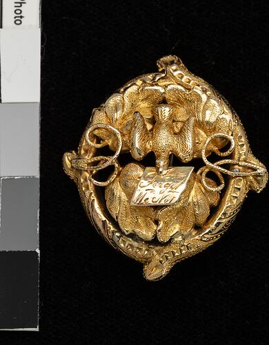 Round gold decorative brooch with bird holding note in its beak.