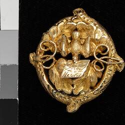 Round gold decorative brooch with bird holding note in its beak.