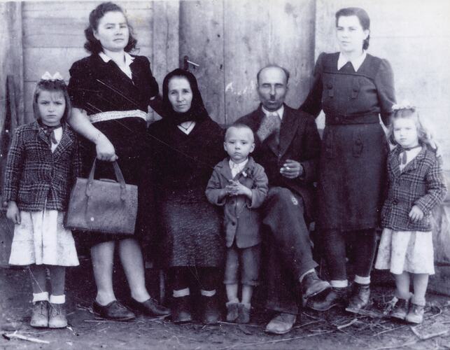Family of seven group portrait. Wooden building behind them.
