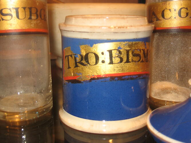 Ceramic cylindrical jar with blue body and white trim. Gold painted label with black text.