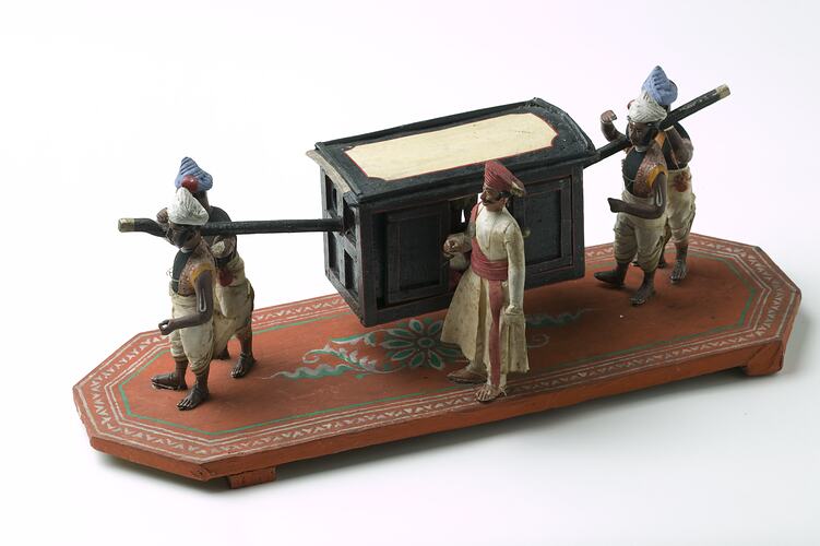 Man in palanquin carried by four men. Another man stands alongside. Wooden base painted orange, green, white.