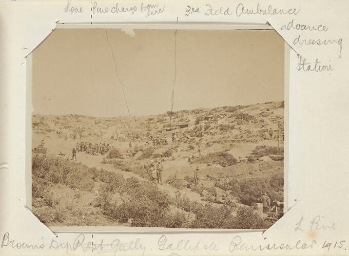 Large group of servicemen scattered across a sloping, scrub covered landscape.