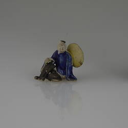 Miniature seated figurine wearing blue top and black pants. Has wide brimmed conical hat.