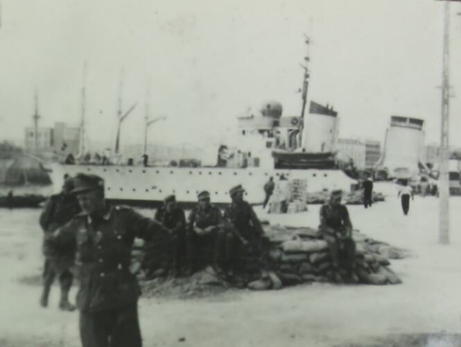 Group of uniformed soldiers sitting on or standing near a pile of sandbags, ship in the background.