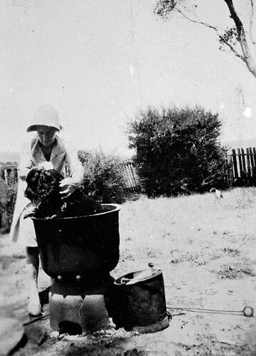 Woman wearing hat washing dog outdoors in copper bath.