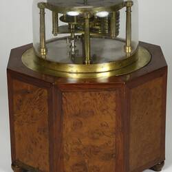 Brass clock under glass dome, supported by wooden octagonal box with door. Front view.