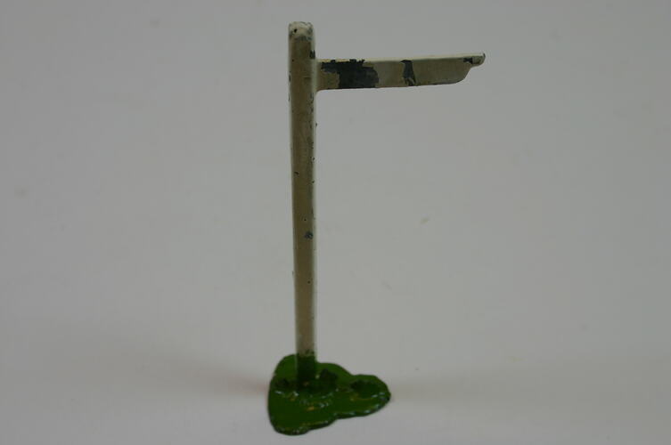 Toy metal signpost. White post and flag. Green grass stand at base.