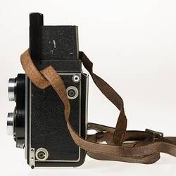 Metal twin lens reflex camera. Body covered in black leather. Leather carry strap. Left profile.