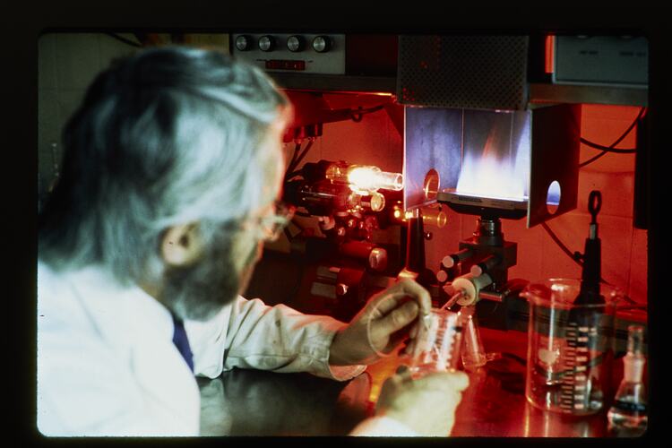 Man sits at red-lit desk with equipment. Holds a glass beaker.