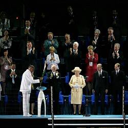 John Landy in white tracksuit placing Olympic baton in its stand as onlookers watch including HRH Queen Elizab