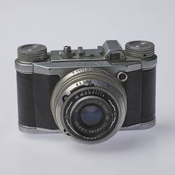 Black and silver camera. Central lens at front and two dials on top.