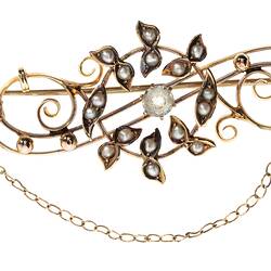 Central gold circle of seed pearls overlaying gold scroll with three paste stones. Fine chain and pin at right.