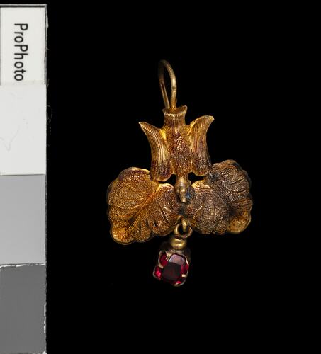 Gold pendant earring with downward facing bird, two leaves and single red hanging charm.