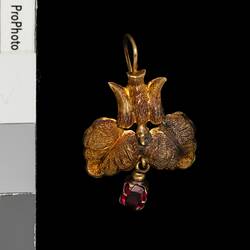 Gold pendant earring with downward facing bird, two leaves and single red hanging charm.