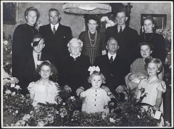 Portrait of nine adults sitting and standing indoors. Three children at front. Seated woman at right holds a baby. Flowers in foreground.