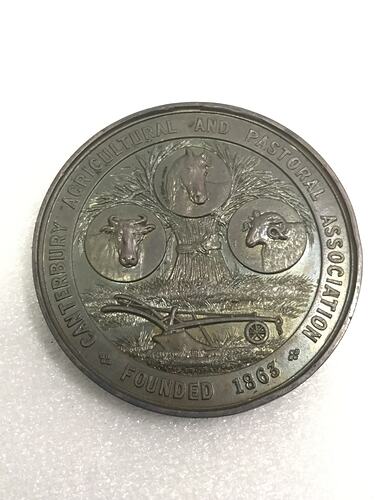 Circular silver medal with a cow, horse and sheep above a sheaf of wheat. Plough below. Text around.