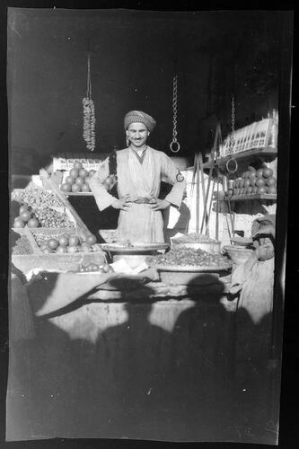 Turbaned man standing, arms akimbo, standing behind his food stall. Trays of produce surround him.