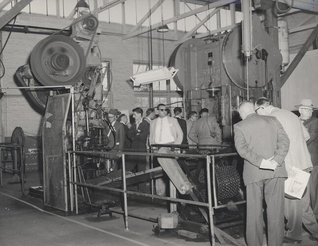 A large group of men on a factory tour inspecting a chain making machine.