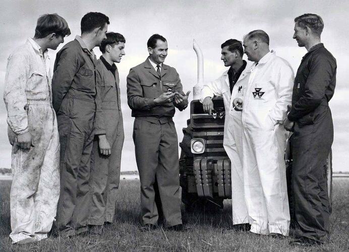 Group of men stand beside tractors in field.