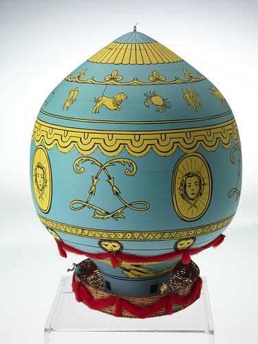 Blue wooden balloon model with yellow patterns. Basket base holds figurines. Side D.