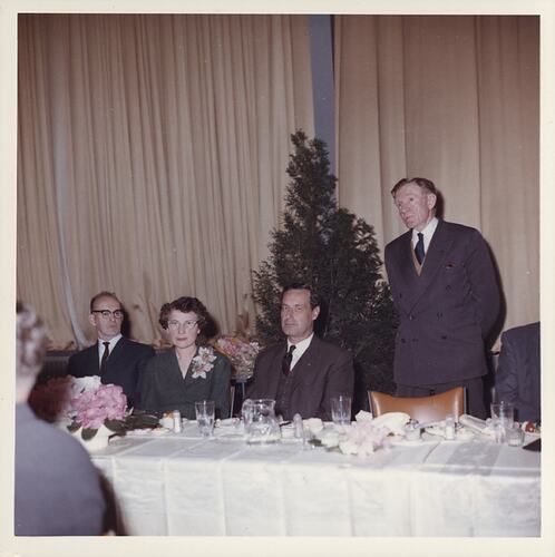 Man speaking at dinner; others look to camera.